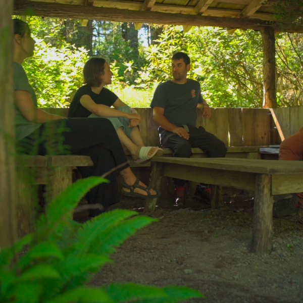 Image gallery pop-up of four individuals sitting and talking inside a wooden shelter in the forest.